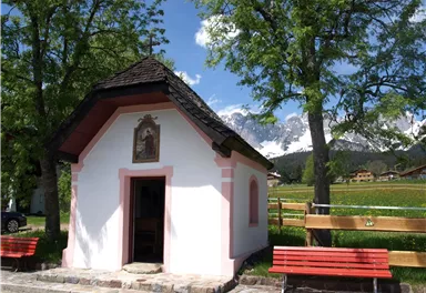 A small chapel with pink accents stands among trees. In the background, mountains and a green meadow can be seen.