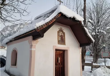Eine kleine Kapelle mit einem Schnee bedeckten Dach. Umgeben von winterlicher Landschaft und schneebedeckten Bäumen.