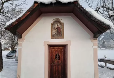 Eine kleine Kapelle mit einem schneebedeckten Dach steht in einer winterlichen Landschaft. Umgeben von schneebedeckten Bäumen und einem ruhigen, friedlichen Ambiente.