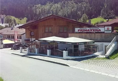 A cozy pizzeria with a wooden facade and outdoor terrace. Surrounded by green mountains and a quiet street.