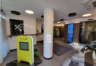 Eine moderne Empfangshalle mit hellen Wänden und einem digitalen Informationsstand. Im Hintergrund gibt es große Fenster und einen Geldautomaten.