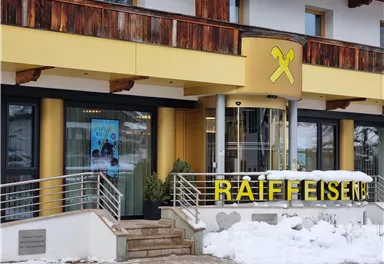 Ein modernes Gebäude der Raiffeisenbank mit einem gelben Schild. Vor dem Eingang liegt Schnee, und es gibt eine Werbetafel an der Wand.