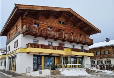 Ein traditionelles Holzhaus mit mehreren Balkonen in den Alpen. Unten befindet sich ein Geschäft mit einem gelben Schild.