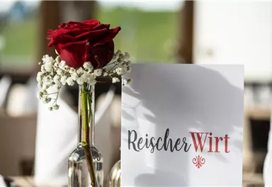 An elegant table decoration with a red rose in a glass vase and a menu card. In the background, a green landscape is visible.