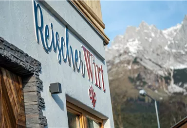 A building with the inscription "ReiseWirt" in a picturesque mountain landscape. The surroundings are mountainous and show snow-capped peaks in the background.