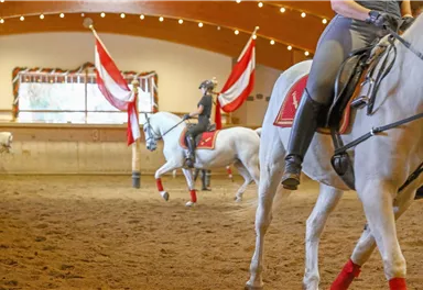 Eine Reithalle mit mehreren Pferden und Reitern. Die Atmosphäre ist festlich mit roten und weißen Fahnen.