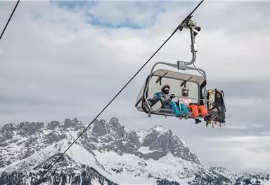 A ski lift cabin with two passengers floats over snow-covered mountains. The sky is cloudy and the landscape looks wintry.