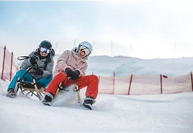 Two people on sleds in the snow. They are having fun and wearing helmets.