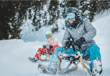 Two people are sitting on sleds and going down a snowy slope. Snow is falling, and the surroundings are surrounded by fir trees.