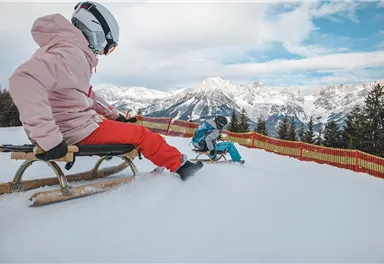 Two children are sledding down a snowy slope. In the background, snow-covered mountains and a clear sky can be seen.