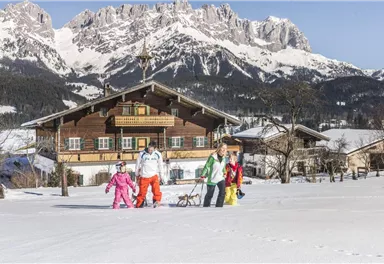 A group of people is pulling sleds through the snow. In the background, snow-covered mountains and a traditional mountain hut can be seen.