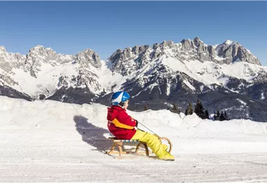A child is sitting on a sled in the snow. In the background, majestic mountains and a clear blue sky can be seen.