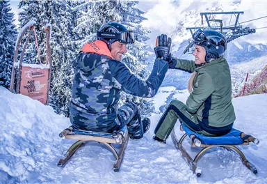 Two people are sitting on sleds in the snow and giving each other a high-five. In the background, there are snow-covered trees and a ski lift.