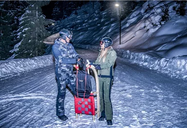 A couple stands on a snow-covered path at night. They are wearing helmets and getting ready to ride a sled.
