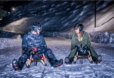 Two people are sitting on sleds in the snow at night. The surroundings are covered in snow and illuminated by a gentle light.