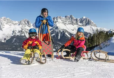 A cheerful group of children is sitting on sleds in the snow. In the background, snow-covered mountains and a bright blue sky can be seen.