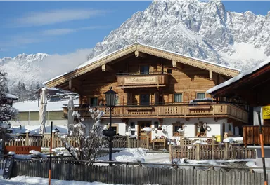 A traditional, wooden-clad chalet in a snowy mountain landscape. In the background, impressive mountains and a clear blue sky can be seen.