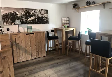 A cozy room with wooden furniture and bar stools. A picture of mountains hangs on the wall, and there is a small workspace by the window.