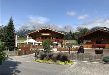An idyllic chalet in the mountains with cozy terraces. The surroundings are green with a view of high mountains and a clear sky.