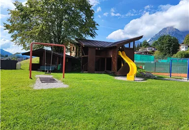 Ein Spielplatz mit einer Rutsche und Schaukeln inmitten einer grünen Wiese. Im Hintergrund sind Bäume und Berge sichtbar.
