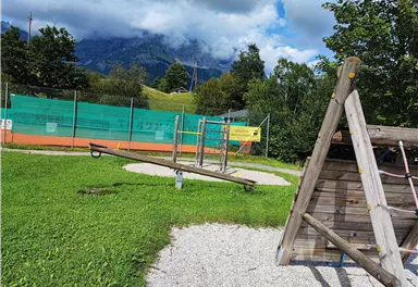 Ein Spielplatz mit einer Wippe und einem Holzgerüst. Im Hintergrund sind Tennisplätze und Berge zu sehen.