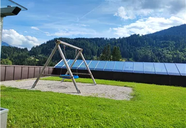 Eine Schaukel auf einem Sandplatz in einem grünen Garten. Im Hintergrund sind Hügel und Bäume zu sehen.