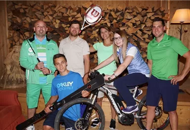 A group of six people is posing in a cozy room with wooden decor. Some are holding sports equipment, including a bicycle and a tennis racket.