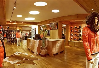A modern sports shop with paneled wooden walls and warm lighting. In the foreground is a stylish mannequin in an orange jacket, surrounded by ski equipment and accessories.