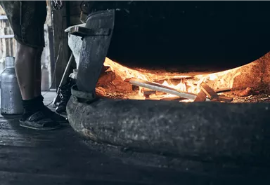 A large cauldron is positioned above a fire that glows orange. You can see the legs of a person and the wooden structure of the floor.
