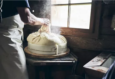 A person prepares fresh cheese in a rural setting. The light softly filters through a window, and the atmosphere is traditional and artisanal.