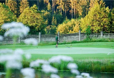 A golfer putts on a green course, surrounded by trees and a blooming meadow. The scene radiates tranquility and a connection to nature.