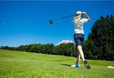 A female golfer hits a ball on a green golf course. The sky is clear and blue, surrounded by trees.