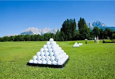A golf course landscape with pyramidal arrangements of golf balls. In the background, there are mountains and a clear blue sky.
