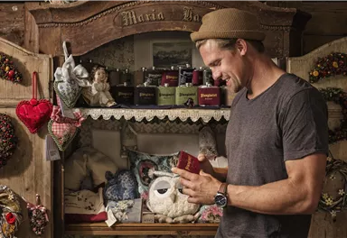 Ein Mann steht an einem rustikalen Regal voller handgefertigter Produkte. Er schaut lächelnd auf ein kleines Geschenk, während im Hintergrund verschiedene Gegenstände und Dekorationen zu sehen sind.