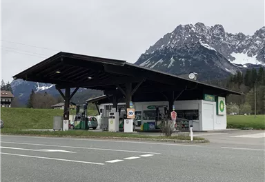 Eine Tankstelle mit einem überdachten Bereich in einer ländlichen Umgebung. Im Hintergrund sind beeindruckende Berge zu sehen.