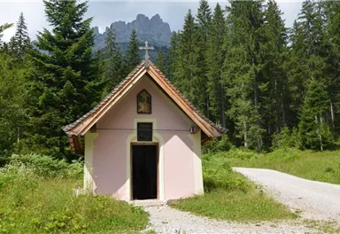A small, pink chapel surrounded by tall trees and green grass. Majestic mountains can be seen in the background.