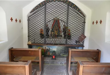 A small chapel with wooden benches and a grille before the altar. In the background, religious figures and flower arrangements can be seen.