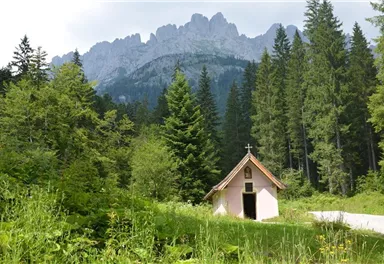 A small chapel stands at the edge of a forest, surrounded by tall trees. Majestic mountains rise in the background.