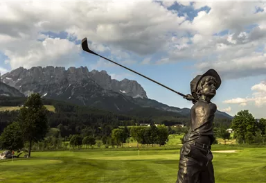 Eine Bronze-Skulptur eines jungen Golfspielers steht auf einem Golfplatz. Im Hintergrund sind majestätische Berge und ein klarer Himmel zu sehen.