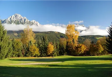 A picturesque landscape with autumn trees and a gentle golf course. In the background, majestic mountains rise beneath a clear sky.
