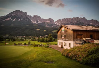 A picturesque golf course and landscape scene with a wooden house in the foreground. In the background, impressive mountains rise under a slightly cloudy sky.