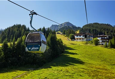 Eine Gondelbahn fährt über eine grüne Wiese in den Bergen. Im Hintergrund sind Wälder und Häuser zu sehen.
