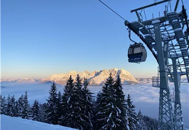 A cable car travels over snow-covered trees in the mountains. In the background, tall mountains are visible under a clear sky.