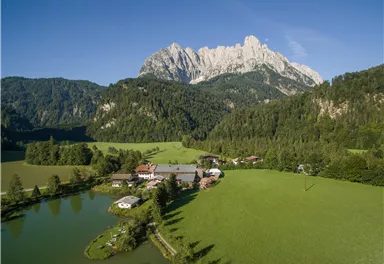 A picturesque landscape with green meadows, a pond, and a farmhouse. In the background, an imposing mountain rises under a blue sky.