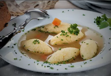 A bowl of broth with fluffy semolina dumplings, garnished with fresh herbs. Next to it is a spoon and some bread.