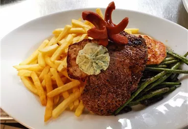 Ein saftiges Steak mit Kräuterbutter, serviert mit Pommes frites, grünen Bohnen und einer Tomatenscheibe. Die Anrichtung ist ansprechend und appetitlich.
