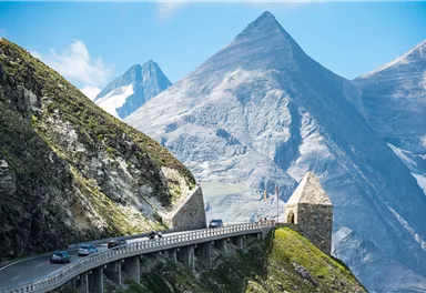 Eine kurvenreiche Straße führt durch eine beeindruckende Berglandschaft. Im Hintergrund ragen majestätische Gipfel in den klaren Himmel.