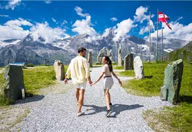 Ein Paar hält Händchen und spaziert auf einem Kiesweg. Im Hintergrund sind beeindruckende Berge und Wolken sichtbar.