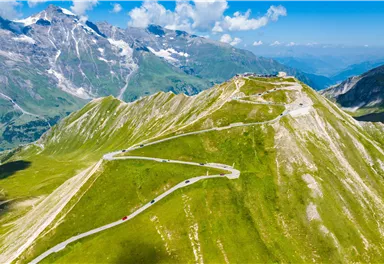 Eine atemberaubende Berglandschaft mit kurvenreicher Straße und grünen Wiesen. Im Hintergrund sind majestätische Gipfel und ein klarer Himmel zu sehen.