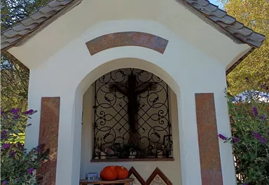 A small chapel with a cross-shaped roof and a wrought iron grid door. On a table in front of the chapel, there is a pumpkin and religious symbols.